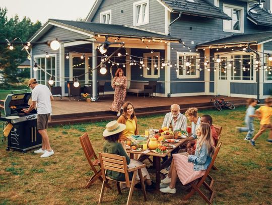 A family is sitting at an outdoor table eating a meal. A man is using the grill and children are running around the backyard.