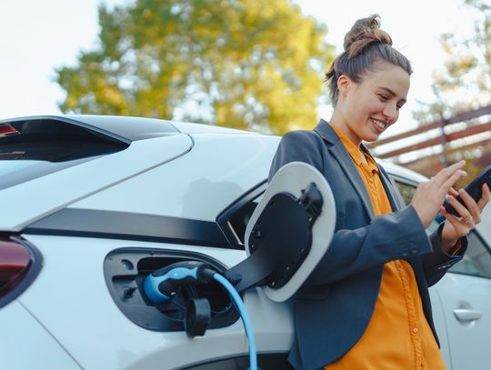 A woman wearing an orange shirt and a blue blazer smiles at her phone while standing next to a white EV that is charging.