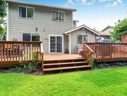 A wooden backyard deck with railings, stairs, outdoor seating, and a grill overlooking a green lawn behind a two-story home.
