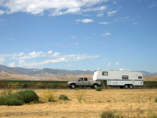 A grey pickup truck towing a large white camper trailer on an empty road with a mountain ridge in the background.