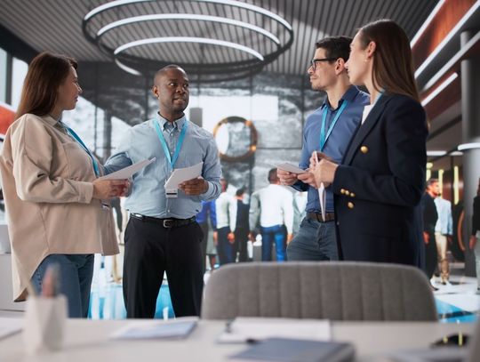 Four professionals wearing teal lanyards holding documents in the middle of a busy trade show floor.