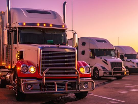 A parking lot contains four semis parked diagonally next to each other. The sunset creates a yellow and purple sky.