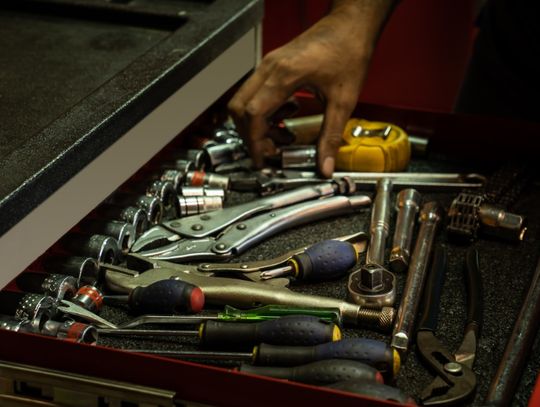 A mechanic reaches into an open tool chest packed with sockets, screwdrivers, wrenches, pliers, and a yellow tape measure.