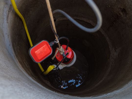 A red pump being lowered into a concrete well with ropes, a yellow hose, black cable, gray looped pipe, and water below.