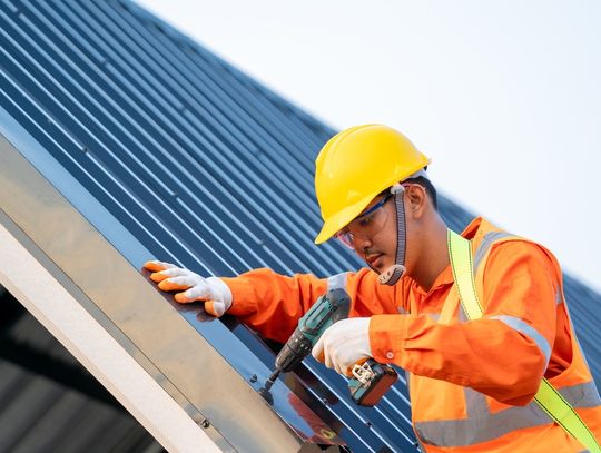A worker wearing a hard hat installing roof panels on a metal roof frame with blue sky in the background.