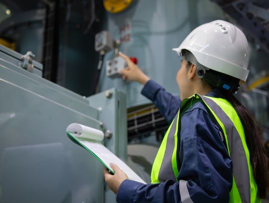 A woman in a white hard hat and green safety vest holds a green clipboard and points toward large equipment.