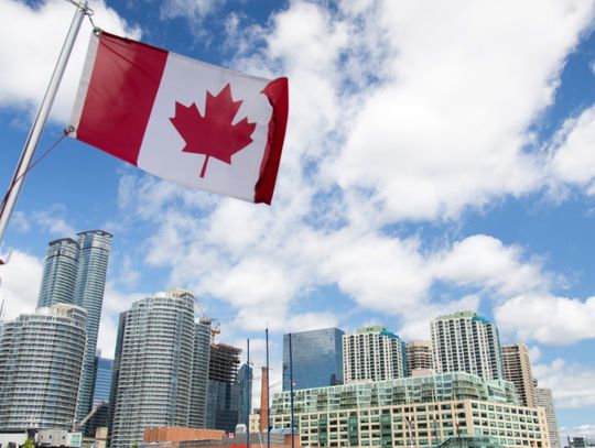 Canadian flag waving above Toronto’s waterfront skyline beneath a blue sky filled with large white clouds.