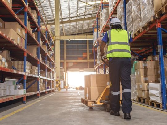 A worker wearing a hard hat and safety vest pulling a pallet truck with boxes through a warehouse aisle.