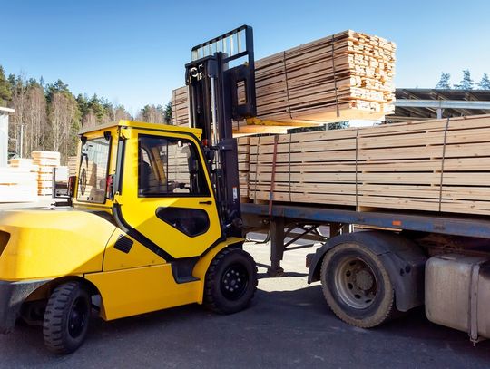 The sun shines down on a yellow forklift as it loads lumber construction materials onto a truck's bed.