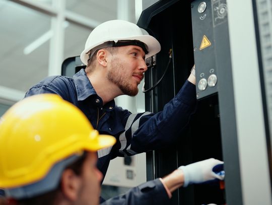 Two engineers wearing hard hats in a brightly lit factory. Both employees make repairs to a large CNC machine.