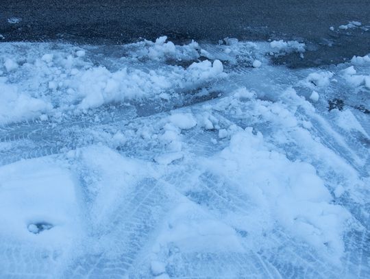 A thin layer of slush covering dark asphalt pavement, marked by tire tracks and some rough, broken-up patches.