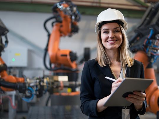 A woman in business clothing and a hard hat stands in front of three manufacturing robots. She holds a tablet in her hand.