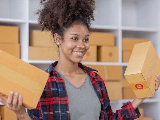 A young Black professional businesswoman is smiling while holding two cardboard boxes in a shipping center.