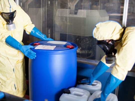 Two people wearing identical protective suits, gloves, and facial coverings stand with chemical storage containers.
