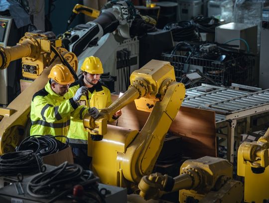 Two engineers wearing yellow hard hats and high-visibility vests inspect a long, yellow robotic arm in a factory.