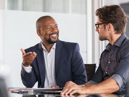 Two men are smiling and talking to one another in a formal business setting. They're both wearing suits.
