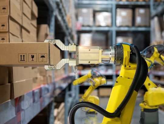 A yellow robotic arm grips a cardboard box beside stacked boxes and shelves in the aisle of a warehouse.