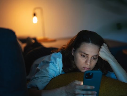 In a dim room, a woman lies on her stomach on a couch. She props her head up with her arm as she looks at her cellphone.