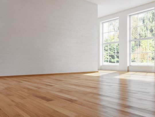 A closeup on the hardwood flooring of house's main room. The walls and ceiling of the room are an off-white color.