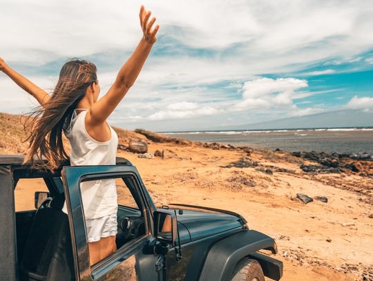 A woman standing up in the passenger seat of a truck with her arms in the air. She is parked in some dusty plains.