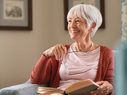 A smiling older woman with short gray hair sits in an armchair holding a book and glasses, relaxing in a cozy living room.