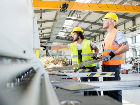 Two employees wearing reflective vests in a manufacturing facility. One person is holding onto sheet metal.