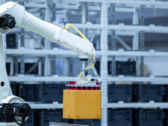 A white and black robotic arm lifts a square cardboard box in the air inside a large warehouse facility.