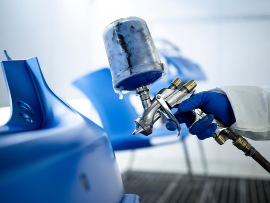 A worker in protective gear uses a spray gun to apply blue paint onto a molded component in an industrial paint booth.