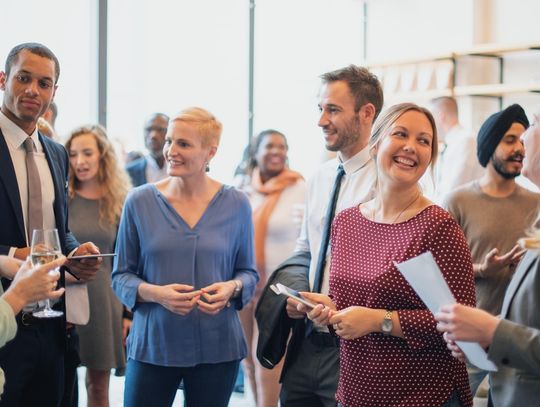 A group of professionals chatting and smiling at a corporate networking event, holding drinks and papers in a bright venue.
