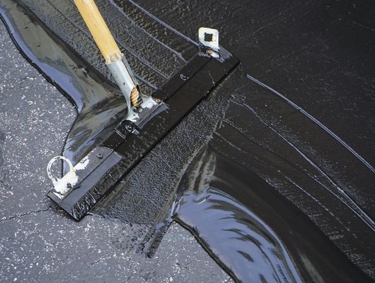 A metal squeegee with a wooden handle spreading glossy black sealant across cracked asphalt pavement.