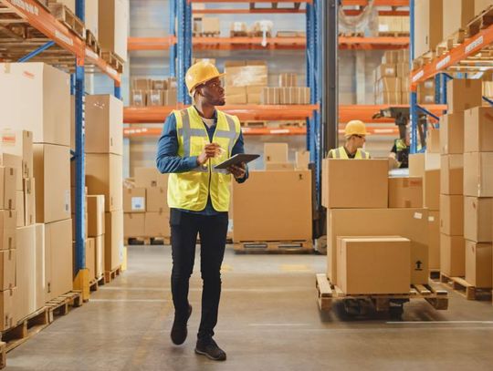 A man in a yellow safety vest and hard hat examining warehouse pallet racking while taking notes on a tablet.