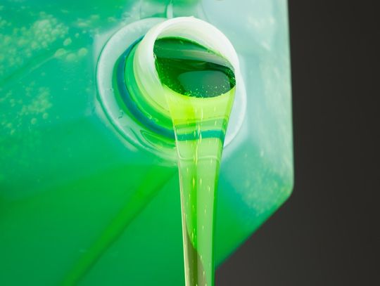 A close-up of a large plastic bottle with bright green liquid detergent pouring from its spout against a black background.