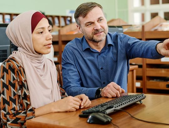 A Caucasian teacher points toward the computer screen while sitting next to a woman wearing a hijab.