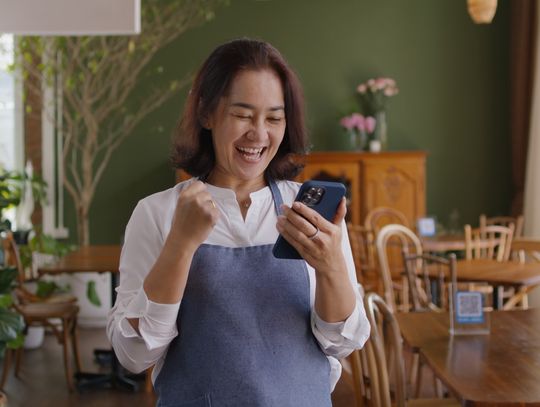 A smiling restaurant owner in an apron celebrates while looking at her phone inside a cozy, plant-filled café dining area.