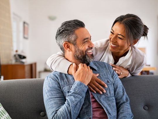 A woman embracing a seated man from behind on a gray couch while they smile at each other in a bright living room.