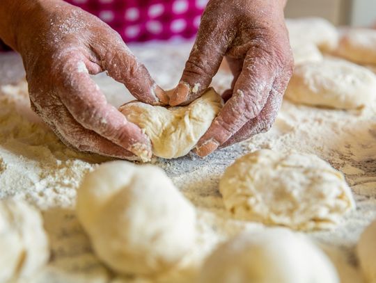 A baker wearing a red checkered apron kneading dough on a floured countertop, surrounded by several other portions of dough.