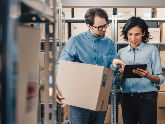 A man and a woman stand in a warehouse looking at a black tablet. The man is holding a cardboard box.