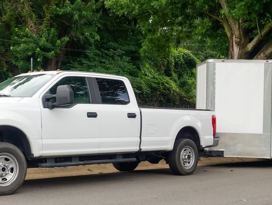 A white trailer attached to a white pickup truck, parked on a street with mature trees in the background.