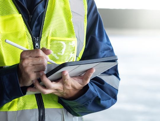 A man in a reflective vest using a digital tablet while inspecting wall-mounted equipment inside a building.