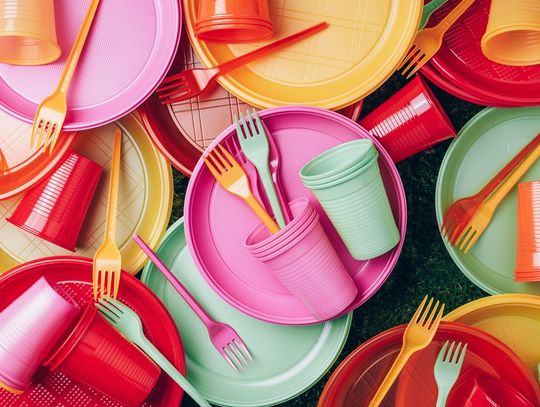 An aerial view shows pink, green, yellow, orange, and red disposable cutlery, plates, and cups scattered across the ground.