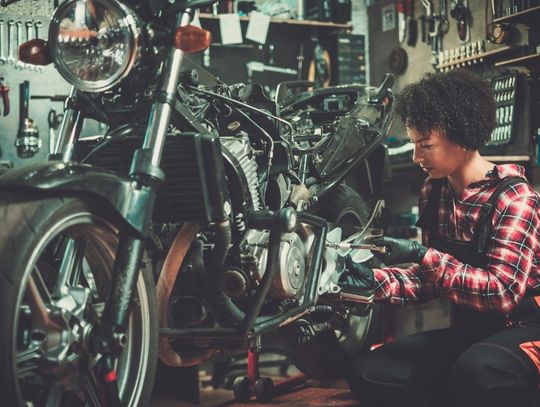 A young woman wearing bib overalls kneels next to a motorcycle and turns a torque wrench. Tools hang on the walls.