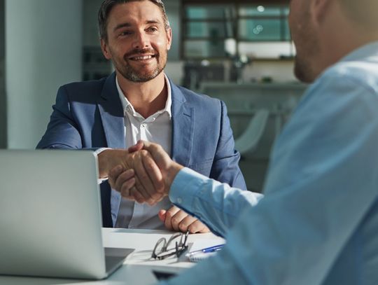 Two professionally dressed employees shaking hands over a large table with a laptop and glasses on it.
