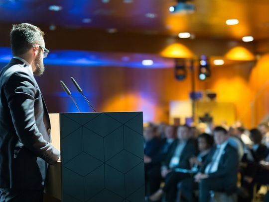A guest speaker standing on the stage at a corporate event. The crowd is full of professionally dressed attendees.