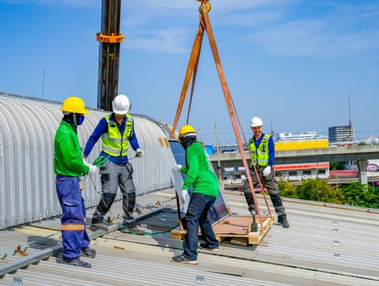 Four construction workers wearing blue-and-green uniforms on a roof using a crane to lift building materials.