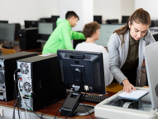 A woman in a gray blazer placing papers on a flatbed scanner in a computer classroom with students seated behind her.