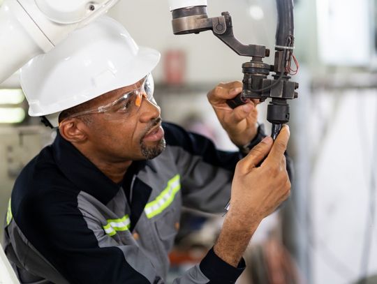 A man wearing a hard hat and safety glasses adjusts two levers on a robotic arm in a brightly lit setting.