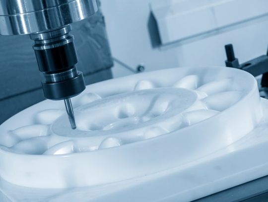 Close-up of a milling bit cutting a smooth white round part on a CNC machine bed in a workshop with cool blue lighting.
