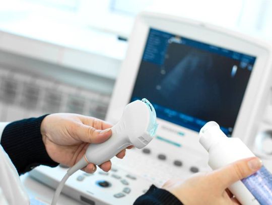 A medical professional applies a blue gel to the end of an ultrasound transducer. The ultrasound machine appears blurry.