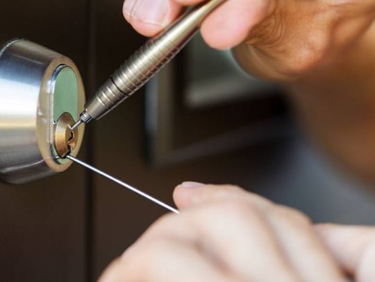 A close-up view shows two hands placing silver lock picking tools into a silver lock on a brown door.