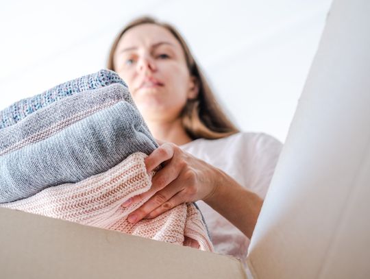 A low-angle view of a woman placing a pile of folded blue and pink sweaters into a brown cardboard box.